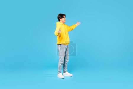 Photo for Positive Korean Teenager Guy Spreading Hands For Hug Meeting Someone Looking Aside Standing Over Blue Studio Background. Boy Advertising Great Offer. Full Length Shot, Side View - Royalty Free Image