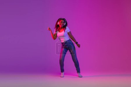 Téléchargez les photos : Sentez le Beat. Femme noire insouciante dans les écouteurs sans fil dansant dans la lumière au néon, jeune femme afro-américaine joyeuse faisant des mouvements énergétiques et s'amusant sur fond violet Studio, Copier l'espace - en image libre de droit