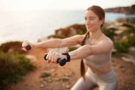 Téléchargez les photos : Énergie positive jeune femme caucasienne en vêtements de sport faire exercice de bras avec haltères, profiter de l'entraînement sur la plage de l'océan, motivant. Soins du corps, soins de santé, fitness et entraînement en plein air - en image libre de droit