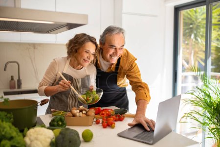 Photo for Happy elderly husband and wife video calling on laptop while cooking dinner in kitchen, spouses browsing internet on computer, preparing healhy vegetarian lunch - Royalty Free Image
