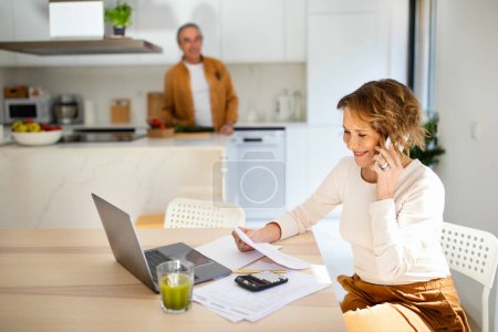 Photo for Happy senior woman talking on smartphone and checking bills when husband preparing dinner at kitchen, selective focus on smiling wife. Solving vacation issues. Utility bills documents - Royalty Free Image
