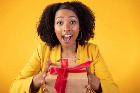 Photo for Excited young african american curly lady holding box with gift and looking at camera with excitement, isolated on yellow background. Surprise, present and congratulations on birthday and holiday - Royalty Free Image