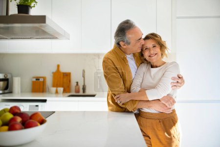 Photo for Happy loving senior spouses in casual outfits embracing, enjoying time together at cozy white kitchen, man kissing his wife, woman smiling at camera, copy space. Seniors lifestyle, family concept - Royalty Free Image