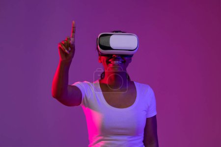 Téléchargez les photos : Femme noire excitée portant un casque Vr jouissant d'une expérience de réalité virtuelle, souriante afro-américaine toucher l'air avec le doigt, debout dans la lumière au néon sur fond violet Studio, Copier l'espace - en image libre de droit