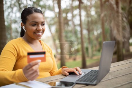 Téléchargez les photos : Joyeux brésilien dame tenant carte de crédit, dactylographier sur ordinateur portable, profiter des ventes en ligne, assis dans le parc de la ville, espace libre. Travail, affaires et freelance à distance, encaissement et achats en ligne - en image libre de droit