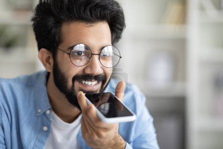 Téléchargez les photos : Gros plan d'un homme indien heureux enregistrant un message vocal sur son smartphone, un jeune homme de l'Est souriant parcourant l'assistant virtuel sur son téléphone portable tout en se reposant à la maison, profitant des technologies modernes - en image libre de droit