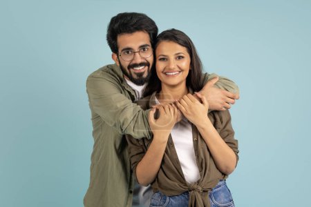 Téléchargez les photos : Beau jeune couple indien posant sur fond bleu studio. Beau bonhomme hindou souriant portant des lunettes embrassant sa jolie petite amie ou sa femme par derrière. Affection, amour - en image libre de droit