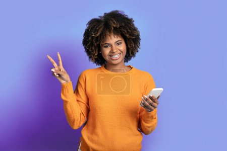 Téléchargez les photos : Joyeux joyeux belle jeune femme noire en pull orange aux cheveux touffus en utilisant un smartphone moderne, montrant geste de paix et souriant sur fond de studio violet, espace de copie - en image libre de droit