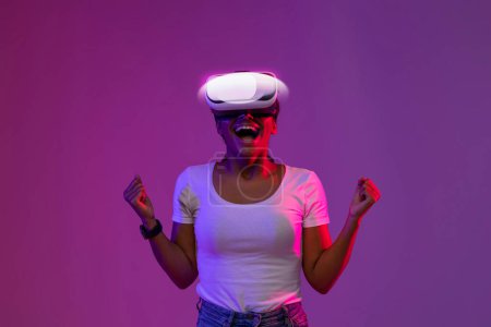Photo for Joyful young black woman wearing glowing VR headset celebrating success while standing in neon light over purple background, happy african american female exploring cyberspace, copy space - Royalty Free Image