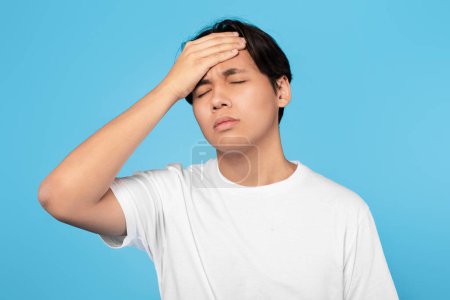 Téléchargez les photos : Malade Japonais adolescent gars ayant fièvre et maux de tête symptôme toucher la tête posant avec les yeux fermés sur fond bleu studio. Malade garçon souffrant de problèmes de santé concept - en image libre de droit