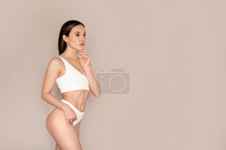 Photo for Beautiful brunette millennial woman posing on beige studio background in comfortable white underwear, touching her hip and chin, looking at copy space. Body treatment, spa concept - Royalty Free Image