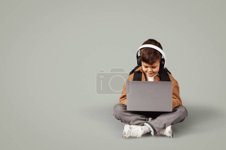 Foto de Ocupado pequeño niño europeo 6 años de edad en mochila casual en auriculares inalámbricos escribiendo en el ordenador portátil, juega juego en línea se sienta en el suelo aislado sobre fondo gris. Lección remotamente, conocimiento, educación - Imagen libre de derechos