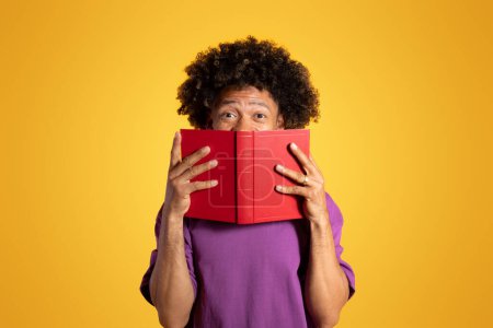 Téléchargez les photos : Joyeux excité adulte afro-américain bouclé homme en violet t-shirt regarde derrière livre isolé sur fond de studio jaune. Lire la littérature, hobby, étude et connaissance - en image libre de droit