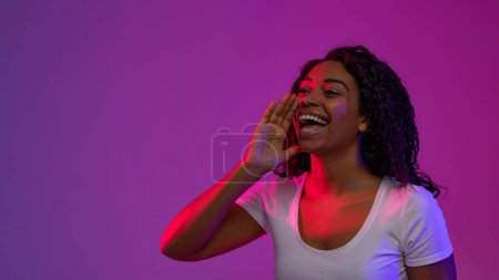 Téléchargez les photos : Jeune femme noire avec la main près de la bouche faisant l'annonce dans la lumière au néon, femme afro-américaine joyeuse partageant des nouvelles ou des informations tout en se tenant debout sur fond violet, Copier l'espace - en image libre de droit