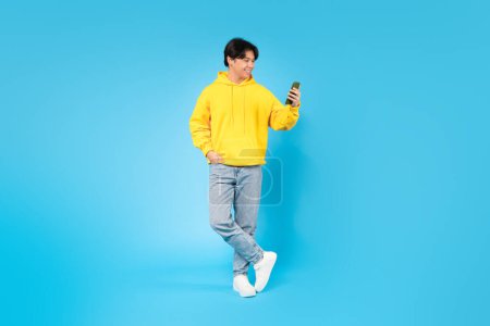 Téléchargez les photos : Korean Teen Boy utilisant un téléphone portable avec une nouvelle application mobile posant debout sur fond bleu. Studio Shot Of Cheerful Guy Texting And Scrolling In Social Media Application. Concept de loisirs numériques - en image libre de droit