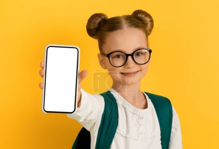 Photo for Study App. Cute little schoolgirl showing blank smartphone with white screen at camera while standing on yellow studio background, smiling female kid recommending educational application, mockup - Royalty Free Image