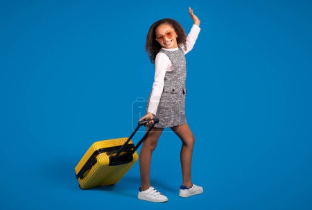 Foto de Feliz adolescente afroamericana chica rizada turista en gafas de sol con maleta disfrutar de viajes aislados en el fondo del estudio azul. Infancia, vacaciones y viajes, estilo de vida activo, vuelo - Imagen libre de derechos