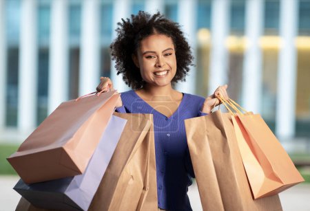 Téléchargez les photos : Joyeux femme noire acheteur publicité offre d'achat tenant des sacs en papier posant debout à l'extérieur près du centre commercial, souriant à la caméra. Grand Saisonnière Sales Publicité Concept - en image libre de droit