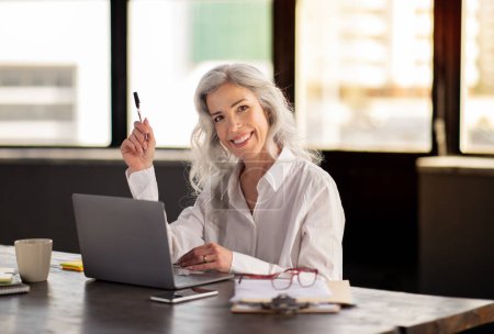 Téléchargez les photos : Entreprise réussie Lady assis près de l'ordinateur portable sur le lieu de travail, souriant à la caméra et tenant stylo posant dans le bureau moderne. Prise de vue d'entrepreneure joyeuse. Concept carrière et emploi - en image libre de droit
