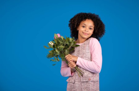 Foto de Feliz chica afroamericana adolescente con ramo de flores celebrando la fiesta, disfrutar de la primavera aislado en el fondo del estudio azul, espacio libre. Saludos de cumpleaños, infancia y vacaciones - Imagen libre de derechos