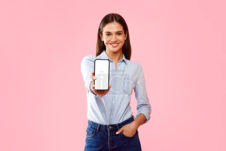 Téléchargez les photos : Publicité App. Heureuse femme caucasienne tenant smartphone avec écran blanc vierge à la main, montrant gadget avec espace vide pour la maquette, bannière, fond rose - en image libre de droit