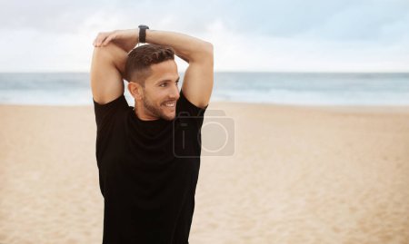 Smiling sporty man doing hand stretching exercises, warming up before training on ocean beach, panorama, free space. Morning workout, body care outdoor