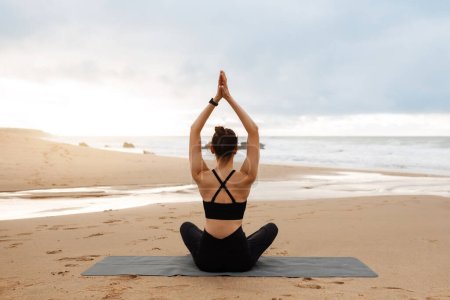 Photo for Outdoor yoga. Woman meditating with hands clasped above head, sitting in lotus position on fitness mat on beach, practicing meditation near ocean, back view - Royalty Free Image