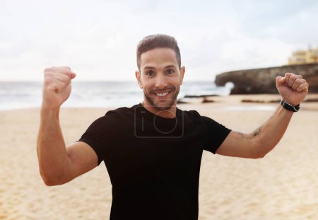 Photo for Excited strong man in sportswear doing success and victory gesture smiling at camera, enjoying effective workout on ocean beach. Morning workout, body care outdoor - Royalty Free Image