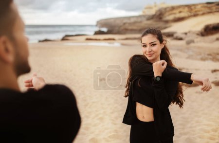 Photo for Excited woman and man doing warm up and stretching for arms in morning on beach near ocean, exercising outdoors. Relationship, health care, training, active lifestyle and sports with trainer - Royalty Free Image