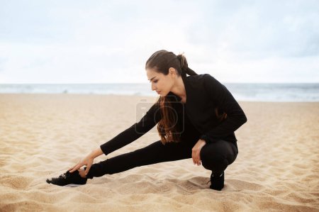 Téléchargez les photos : Femme sportive en entraînement de vêtements de sport sur la plage, étirement des muscles des jambes tout en se préparant à l'exercice près de la mer, échauffement avant le jogging, espace libre - en image libre de droit