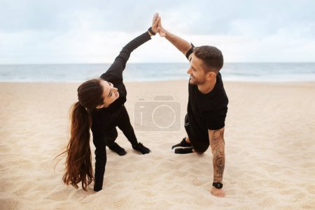 Téléchargez les photos : Joyeux couple en tenue de sport debout dans la planche latérale et donnant cinq haut, l'entraînement le matin sur la plage de l'océan, à l'extérieur. Relation, soins de santé, formation axée sur les résultats, mode de vie actif - en image libre de droit