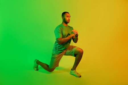 Téléchargez les photos : Entraînement. Afro-Américain Fitness Guy faisant de l'exercice de poussée en vert et jaune néon lumière sur fond de studio. Homme sportif portant des vêtements de sport de formation à la recherche à l'extérieur. Vue latérale - en image libre de droit