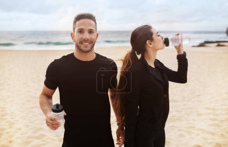Photo for Young fitness couple in sportswear taking break while working out on ocean beach, having break and drinking water, man looking and smiling at camera - Royalty Free Image