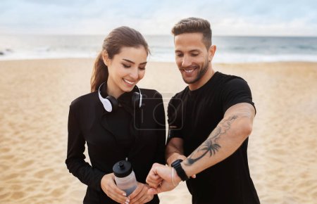 Téléchargez les photos : Heureux sportif homme et femme se reposant après avoir fait du jogging au bord de la mer, vérifier smartwatch fitness tracker, profiter de l'entraînement à l'extérieur sur la plage, espace libre - en image libre de droit