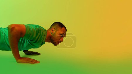 Photo for Fit Black Guy Exercising Standing In Plank Position Looking Aside In Studio, Lit With Green And Yellow Neon Light. Bodybuilding, Fitness Workout Concept. Panorama With Copy Space - Royalty Free Image
