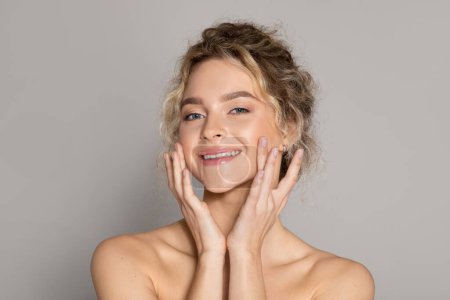 Photo for Facial skincare. Happy beautiful lady touching face, moisturizing smooth skin and smiling at camera, posing over grey studio background. Beauty routine and pampering concept - Royalty Free Image