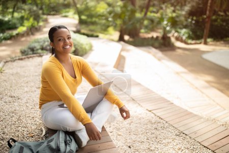 Photo for Happy brazilian lady student sitting on bench with laptop, resting outdoors in park, looking and smiling at camera. Female enjoying online education and distance learning, copy space - Royalty Free Image