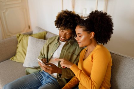 Téléchargez les photos : Femme afro-américaine joyeuse pointant du doigt sur Smartphone naviguant sur Internet avec son mari assis sur le canapé à la maison. Couple utilisant le téléphone et la publicité Nouvelle application mobile. Concept Gadgets - en image libre de droit
