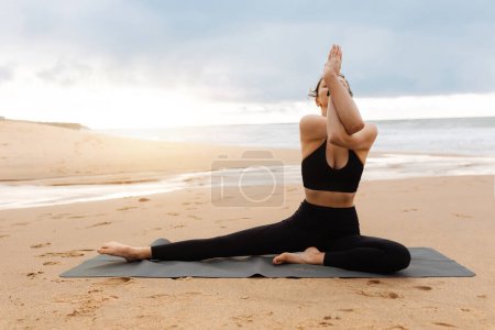 Foto de Yoga matutino al aire libre. Mujer joven en entrenamiento de ropa deportiva al aire libre, mujer atlética sentada en una colchoneta de fitness en yoga asana, haciendo ejercicio en la playa cerca del océano, espacio libre - Imagen libre de derechos