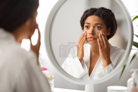 Foto de Dull Skin Concept. Mujer negra molesta mirando el espejo y la cara conmovedora, mujer afroamericana decepcionada sentada en el tocador, revisando círculos oscuros debajo de los ojos y las arrugas - Imagen libre de derechos