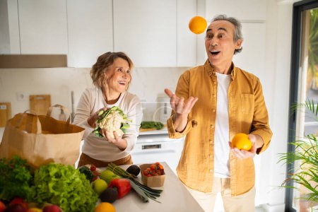 Téléchargez les photos : Homme âgé européen excité jonglant avec des oranges dans un intérieur de cuisine confortable, les conjoints s'amusent tout en déballant des légumes du sac en papier d'épicerie - en image libre de droit