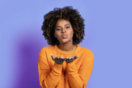 Photo for Beautiful cheerful young african american woman with bushy curly hair outstretching hands towards camera and showing fly kiss, female expressing love and affection on purple studio background - Royalty Free Image