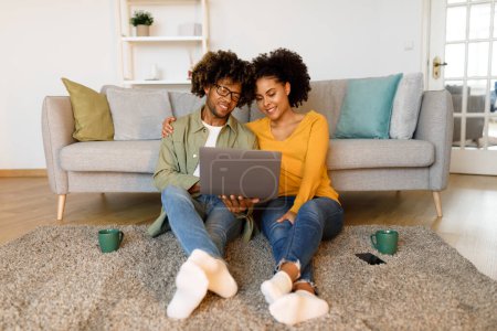 Téléchargez les photos : Carrière freelance. Heureux couple afro-américain en utilisant ordinateur portable assis sur le sol à la maison. Jeunes conjoints surfant sur Internet via les gadgets passer le week-end dans le salon moderne intérieur - en image libre de droit