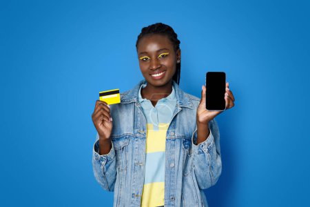Téléchargez les photos : Heureuse jeune femme noire souriante et positive portant une tenue décontractée et un maquillage lumineux montrant une carte bancaire jaune en plastique et un smartphone avec écran vide noir, une maquette, un fond bleu, des achats en ligne - en image libre de droit