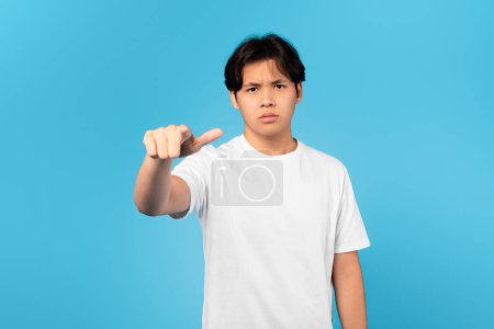 Foto de Mírate. Infeliz coreano adolescente chico señalando dedo y frunciendo el ceño mirando a la cámara de pie posando en el estudio sobre fondo azul. Mala elección y concepto de emociones negativas - Imagen libre de derechos