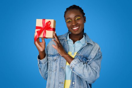 Photo for Happy smiling pretty young african american lady wearing stylish casual outfit and bright makeup going birthday party, holding gift box with red bow, isolated on blue studio background - Royalty Free Image