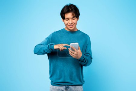 Téléchargez les photos : Nouvelle application. coréen adolescent garçon à l'aide Smartphone défilement médias sociaux Feed in application debout sur bleu studio fond. Guy Jouer à des jeux en ligne Via Gadget. Concept d'offre mobile - en image libre de droit