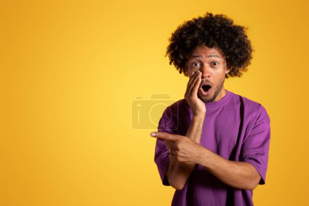 Photo for Shocked adult black curly man presses hand to open mouth, points finger to empty space isolated on yellow studio background. Recommendation, surprise emotions, huge sale, ad and offer - Royalty Free Image