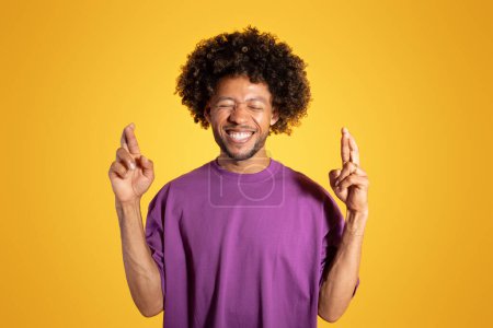 Photo for Glad adult african american curly man in purple t-shirt with closed eyes, crossed fingers, makes a wish, expects to good news on yellow studio background. Dream, ad and offer - Royalty Free Image