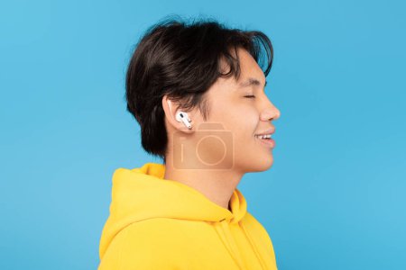 Photo for Side View Portrait Of Asian Teenager Boy Wearing Earbuds Earphones Listening To Music Posing Over Blue Studio Background. Portrait Of Teen Guy Relaxing And Enjoying Song With Eyes Closed - Royalty Free Image